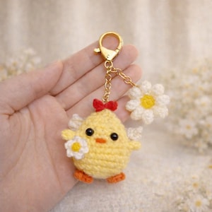 Könnte beinhalten: Ein handgefertigter gelber Häkel-Küken-Schlüsselanhänger mit roter Schleife, orangefarbenen Füßen und weißen Gänseblümchen-Akzenten. Der Schlüsselanhänger hat einen goldenen Verschluss und eine Kette. Das Küken hält eine kleine Gänseblümchen.