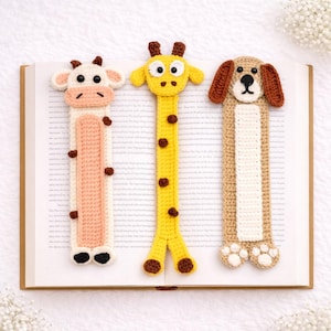 May include: Three handmade crochet bookmarks: a cow, a giraffe, and a dog. The cow bookmark is white and pink, the giraffe is yellow with brown spots, and the dog is tan and white. All are placed on an open book.