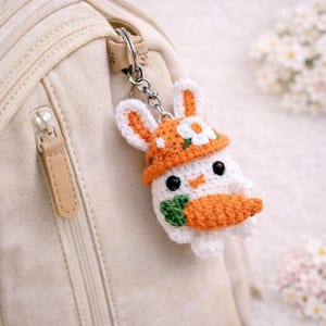 May include: A handmade crochet bunny keychain with an orange hat and carrot. The white bunny has black eyes and a small orange nose. The keychain is attached to a silver metal ring and chain, hanging from a beige backpack.