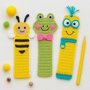 Può includere: Tre segnalibri all'uncinetto fatti a mano: un'ape gialla, una rana verde con un papillon rosa e un personaggio giallo con accenti blu. Una matita gialla è appoggiata accanto ai segnalibri. I segnalibri sono progettati con facce sorridenti.