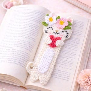 May include: A handmade crochet cat bookmark with a floral crown and a red heart. The bookmark is cream-colored with a white belly and is placed in an open book. The cat's face has embroidered details.