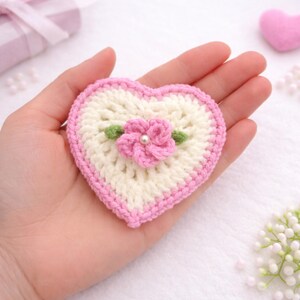 May include: A handmade crochet heart with a pink border and a cream-colored center. A pink flower with green leaves and a pearl center is in the middle. The heart is approximately 3 inches wide.