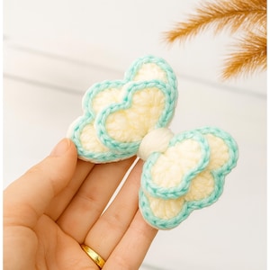 Pode incluir: Um laço de cabelo de crochê feito à mão em forma de borboleta. O laço apresenta um centro amarelo claro com uma borda turquesa. O laço é segurado em uma mão, mostrando o intrincado trabalho de costura e a combinação de cores.