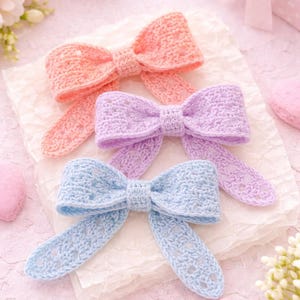 May include: Three pastel-coloured, handmade crochet bows: peach, lavender, and light blue. The bows are arranged on textured paper, with heart-shaped decorations and flowers in the background. A charming, delicate craft.