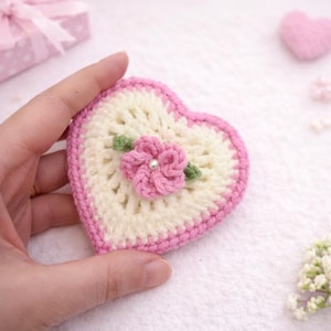 May include: A handmade crochet heart in cream, outlined in pink, with a pink flower and green leaves. The heart is held in a hand, with a soft, blurred background.