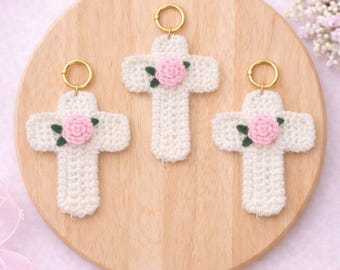 May include: Three white crochet crosses with pink rose accents and gold key rings are displayed on a wooden surface. The crosses are arranged in a row, each featuring a detailed floral design. The background is a soft pink, with floral accents.
