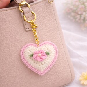 May include: A handmade crochet heart keychain with a pink and white design, featuring a pink flower with a pearl center and green leaves. The keychain is attached to a gold chain and a light pink purse.