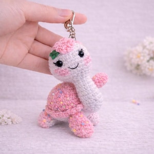 Può includere: Un portachiavi a forma di tartaruga all'uncinetto fatto a mano. La tartaruga ha testa e collo bianchi, un guscio rosa con macchie colorate e arti rosa. Presenta occhi neri, guance rosee e una bocca sorridente. Un anello portachiavi argentato è attaccato alla testa della tartaruga.