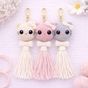 May include: Three handmade crochet cat keyrings in cream, pink, and grey. Each cat has a bow, black eyes, and a tassel. The keyrings are attached to gold-coloured clasps. A charming and whimsical accessory.