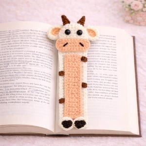 Può includere: Un segnalibro all'uncinetto fatto a mano a forma di mucca, appoggiato su un libro aperto. Il segnalibro presenta un corpo bianco, un viso e un centro color pesca e dettagli marroni. La mucca ha occhi neri e piccole corna.