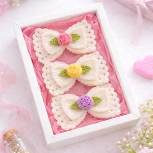 May include: A set of three cream crochet bows with a scalloped design, each featuring a colourful flower and green leaf, presented in a white box on pink tissue paper. The flowers are pink, yellow, and purple.