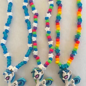 May include: Three colorful beaded necklaces with pony pendants. The necklaces feature star-shaped and round beads in blue, rainbow, and other colors. Each pendant is a white pony with blue hair and sunglasses.