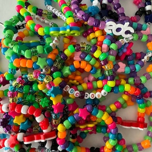 Pode incluir: Um close-up de um monte de pulseiras de contas coloridas com várias letras e símbolos. As pulseiras são feitas de pequenas contas redondas em uma variedade de cores, incluindo vermelho, laranja, amarelo, verde, azul, roxo e rosa. Algumas das pulseiras têm letras que soletram palavras como "EDC 2024", "Wasteland" e "Basspod".