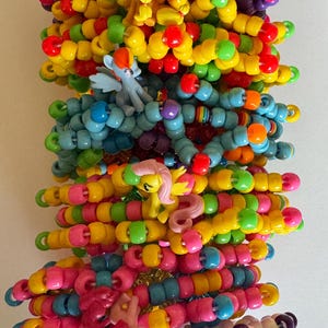 May include: A stack of colorful beaded bracelets with small plastic pony figures attached. The bracelets are made with various colors of beads, including yellow, pink, blue, green, and purple.