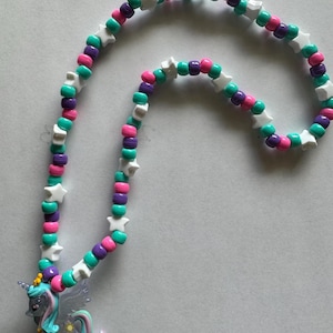 May include: A necklace featuring a series of round beads in shades of teal, purple, and pink, interspersed with white star-shaped beads. A translucent, winged unicorn pendant with a teal mane and tail hangs from the bottom of the beaded strand.