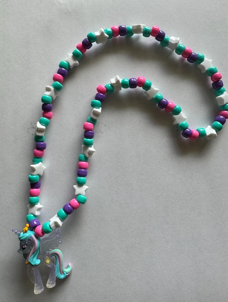 May include: A necklace featuring a series of round beads in shades of teal, purple, and pink, interspersed with white star-shaped beads. A translucent, winged unicorn pendant with a teal mane and tail hangs from the bottom of the beaded strand.