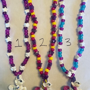 May include: Three beaded necklaces labelled 1, 2, and 3. Each necklace features colourful plastic pony charms and a pattern of round and star-shaped beads in purple, yellow, white, pink, and blue. The charms depict white ponies with colourful manes.