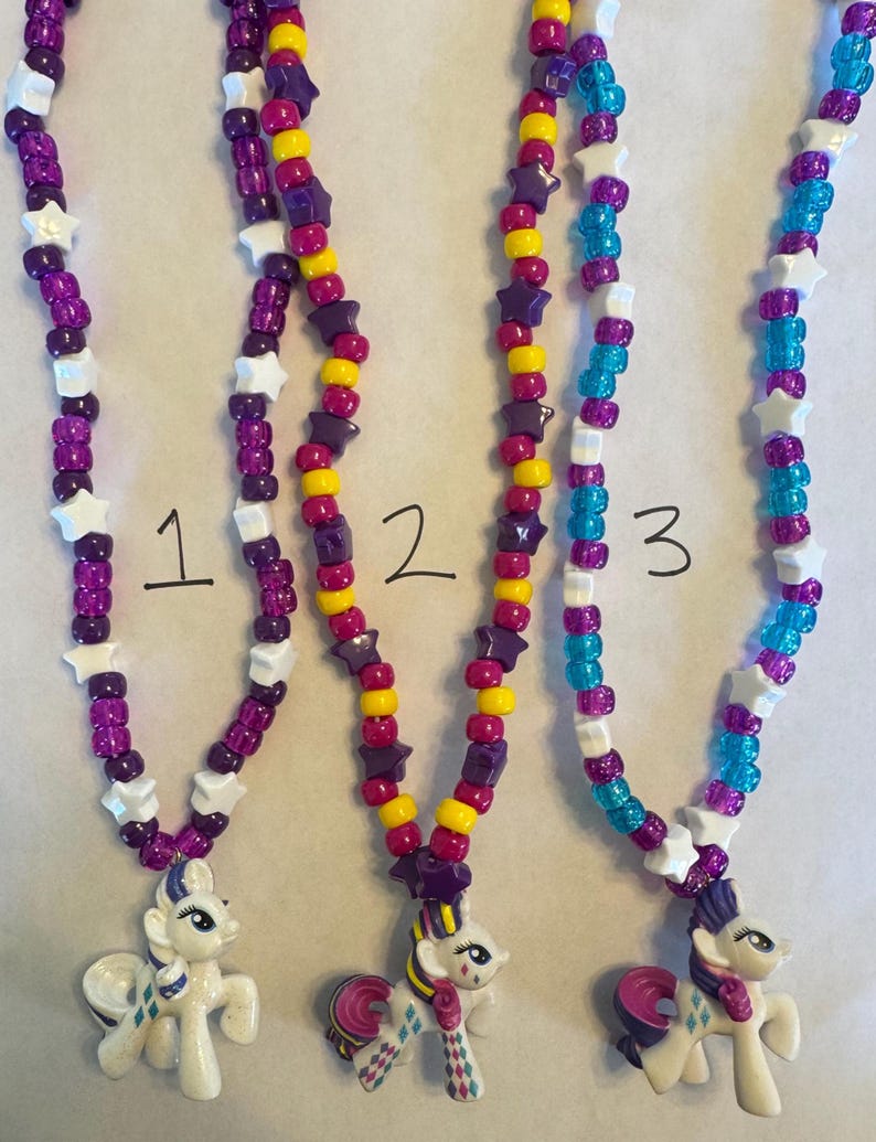 May include: Three beaded necklaces labelled 1, 2, and 3. Each necklace features colourful plastic pony charms and a pattern of round and star-shaped beads in purple, yellow, white, pink, and blue. The charms depict white ponies with colourful manes.