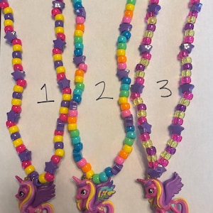 May include: Three necklaces labelled 1, 2, and 3, each featuring a pink unicorn pendant with purple wings and yellow hair. The strands are made of colourful plastic pony beads in shades of yellow, orange, pink, blue, and purple, interspersed with purple star-shaped beads.