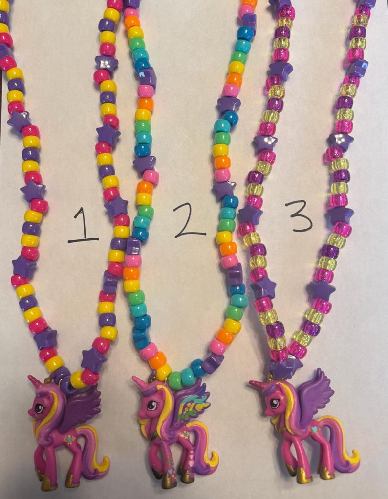 May include: Three necklaces labelled 1, 2, and 3, each featuring a pink unicorn pendant with purple wings and yellow hair. The strands are made of colourful plastic pony beads in shades of yellow, orange, pink, blue, and purple, interspersed with purple star-shaped beads.