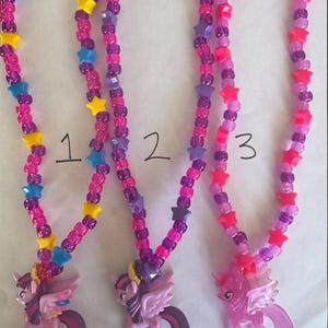 May include: Three necklaces featuring pony pendants and colourful plastic beads. Each necklace is strung with pink, purple, and translucent beads interspersed with yellow, blue, and pink star-shaped beads. The pendants are purple and pink pony figures.