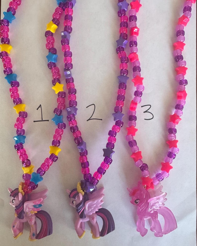 May include: Three necklaces featuring pony pendants and colourful plastic beads. Each necklace is strung with pink, purple, and translucent beads interspersed with yellow, blue, and pink star-shaped beads. The pendants are purple and pink pony figures.