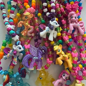 May include: A collection of colourful beaded necklaces featuring various My Little Pony character charms. The necklaces are made with a mix of translucent, glittery, and solid-coloured plastic beads in shades of pink, yellow, blue, green, and purple.