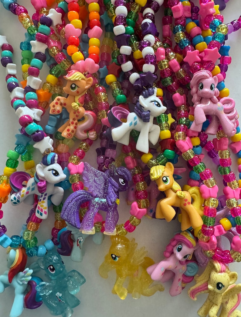 May include: A collection of colourful beaded necklaces featuring various My Little Pony character charms. The necklaces are made with a mix of translucent, glittery, and solid-coloured plastic beads in shades of pink, yellow, blue, green, and purple.
