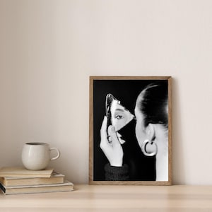 Op de afbeelding: Zwart-wit ingelijste kunstafdruk met een persoon die een scherf van een gebroken spiegel vasthoudt, waarin een oog wordt weerspiegeld. Het kunstwerk heeft een zwarte achtergrond en een houten lijst. Een koffiekopje en boeken staan op een houten oppervlak.