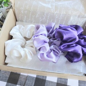 May include: Three satin scrunchies in a gift box. One scrunchie is white, one is light purple, and one is dark purple.