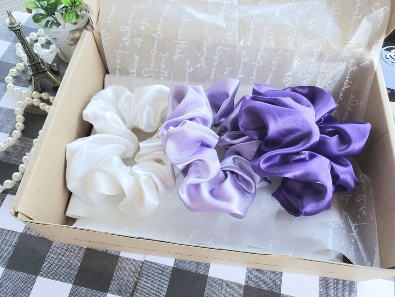 May include: Three satin scrunchies in a gift box. One scrunchie is white, one is light purple, and one is dark purple.