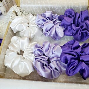 May include: A set of six satin scrunchies in a gift box. The scrunchies are in three colors: white, light purple, and dark purple.