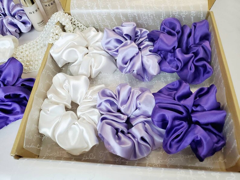 May include: A set of six satin scrunchies in a gift box. The scrunchies are in three colors: white, light purple, and dark purple.