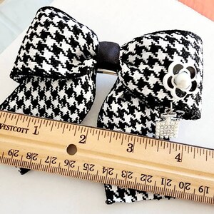 May include: Black and white houndstooth bow with a silver flower and rhinestone charm. The bow measures approximately 4 inches wide.