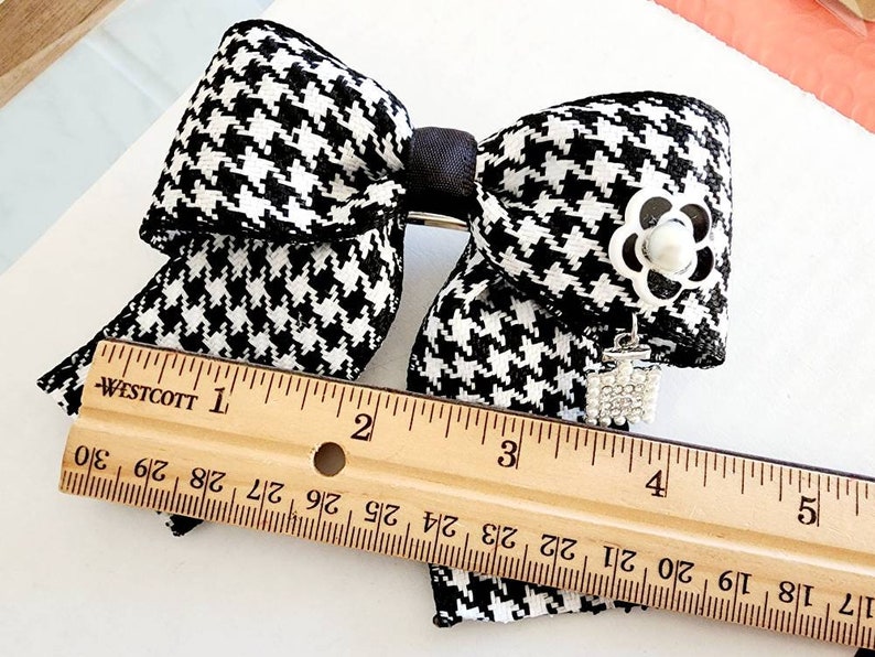 May include: Black and white houndstooth bow with a silver flower and rhinestone charm. The bow measures approximately 4 inches wide.