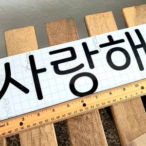 Könnte beinhalten: Ein weißer Aufkleber mit schwarzer Schrift, auf dem "사랑해" steht, was im Koreanischen "Ich liebe dich" bedeutet.