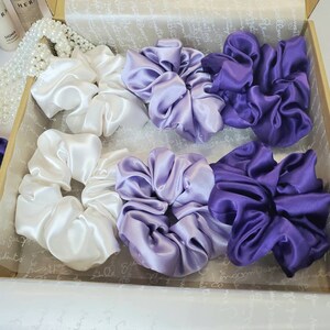 May include: A set of six satin scrunchies in a box. There are two white scrunchies, two light purple scrunchies, and two dark purple scrunchies.