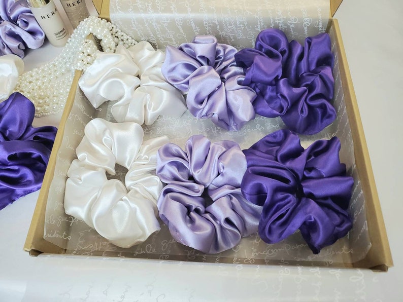 May include: A set of six satin scrunchies in a box. There are two white scrunchies, two light purple scrunchies, and two dark purple scrunchies.