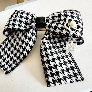 May include: Black and white houndstooth bow with a white pearl and black flower accent. The bow has a silver charm with rhinestones hanging from it.