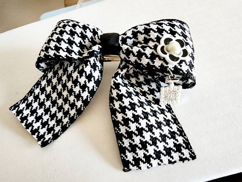 May include: Black and white houndstooth bow with a white pearl and black flower accent. The bow has a silver charm with rhinestones hanging from it.