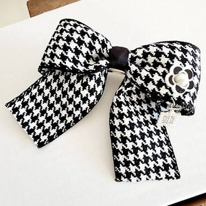 May include: A black and white houndstooth bow with a pearl-like button in the center. The bow is attached to a silver clip.