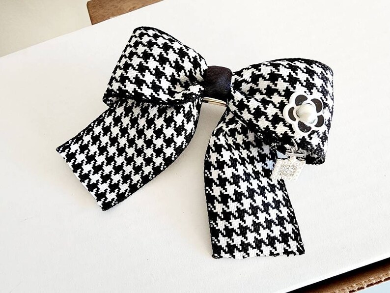 May include: A black and white houndstooth bow with a pearl-like button in the center. The bow is attached to a silver clip.