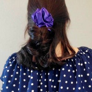 May include: A woman with long, dark brown hair is wearing a navy blue and white polka dot blouse. Her hair is pulled back with a purple satin scrunchie.
