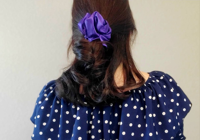 May include: A woman with long, dark brown hair is wearing a navy blue and white polka dot blouse. Her hair is pulled back with a purple satin scrunchie.