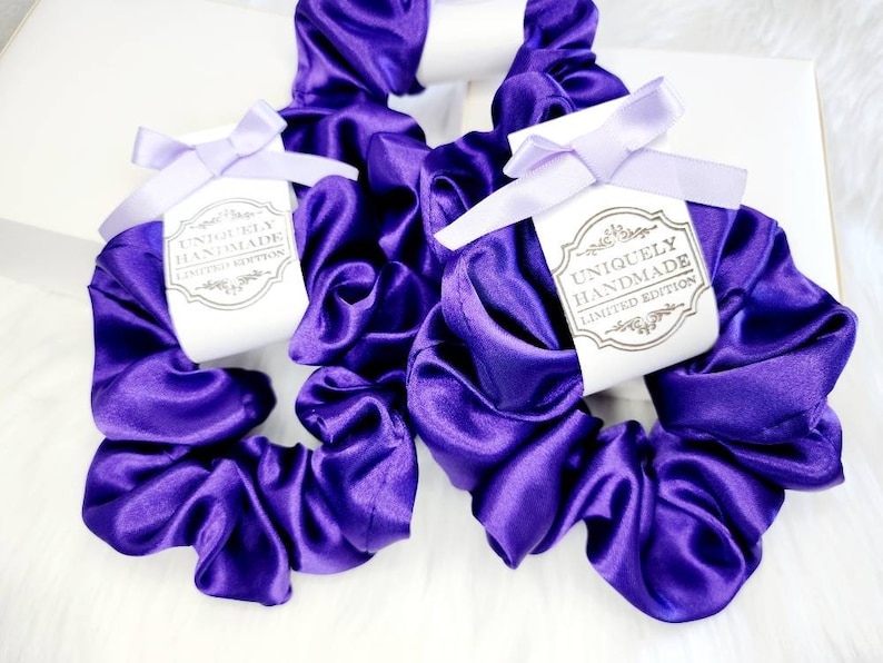 May include: Two purple satin scrunchies with white labels that say "Uniquely Handmade Limited Edition".