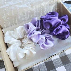 May include: Three satin scrunchies in a gift box. One scrunchie is white, one is light purple, and one is dark purple.