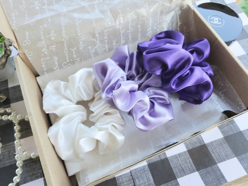 May include: Three satin scrunchies in a gift box. One scrunchie is white, one is light purple, and one is dark purple.