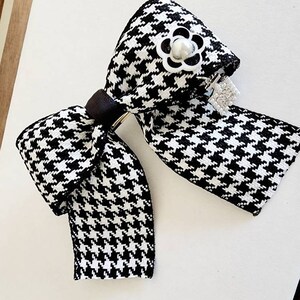 May include: A black and white houndstooth bow with a pearl and silver flower accent. The bow is attached to a silver clip.