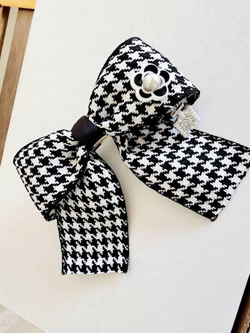 May include: A black and white houndstooth bow with a pearl and silver flower accent. The bow is attached to a silver clip.