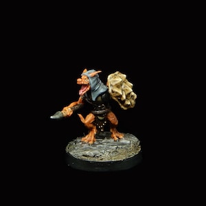 Puede incluir: Figura de fantasía en miniatura de una criatura con pelaje naranja, una capucha azul y un escudo con forma de calavera. Sostiene un pequeño cuchillo y se encuentra sobre una base texturizada. La figura está sobre un fondo negro.